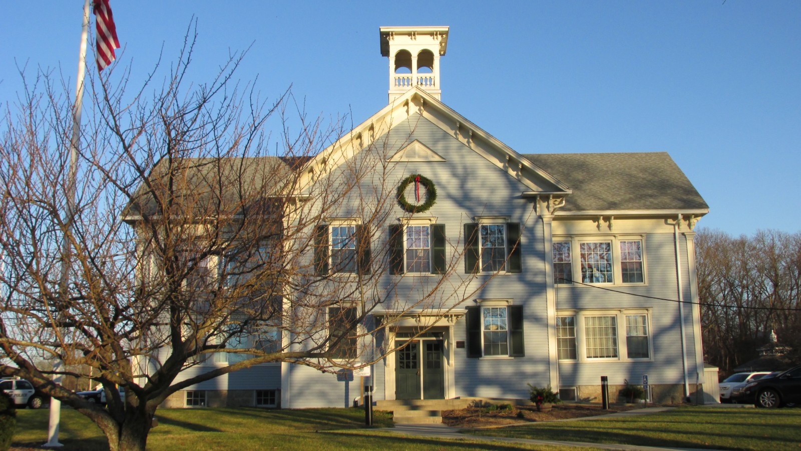 The Parting Ways Building in Acushnet, a two-story pale blue clapboard building with white trim, a cupola, green doors and a wreath in the gable, with a flagpole and a bare tree on the lawn in front
