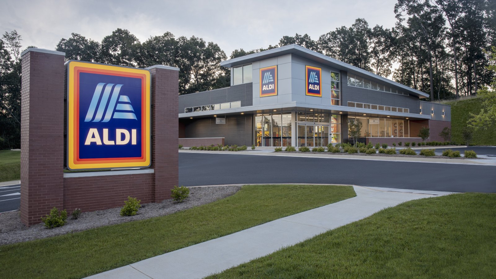 An ALDI store with a tall ALDI sign on a brick monument out front, a gray and glass storefront with two ALDI logos, and a curving drive and lawn at dusk