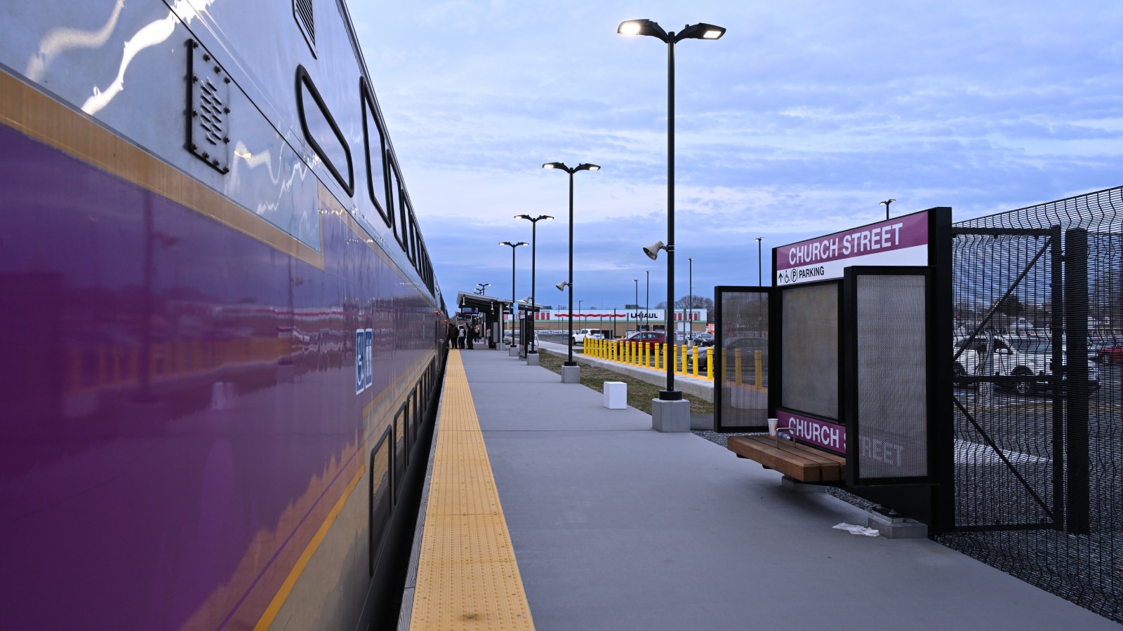 A purple MBTA Commuter Rail coach stopped at the Church Street station platform in New Bedford, with a Church Street parking sign, lamp posts and the station parking lot behind a fence.