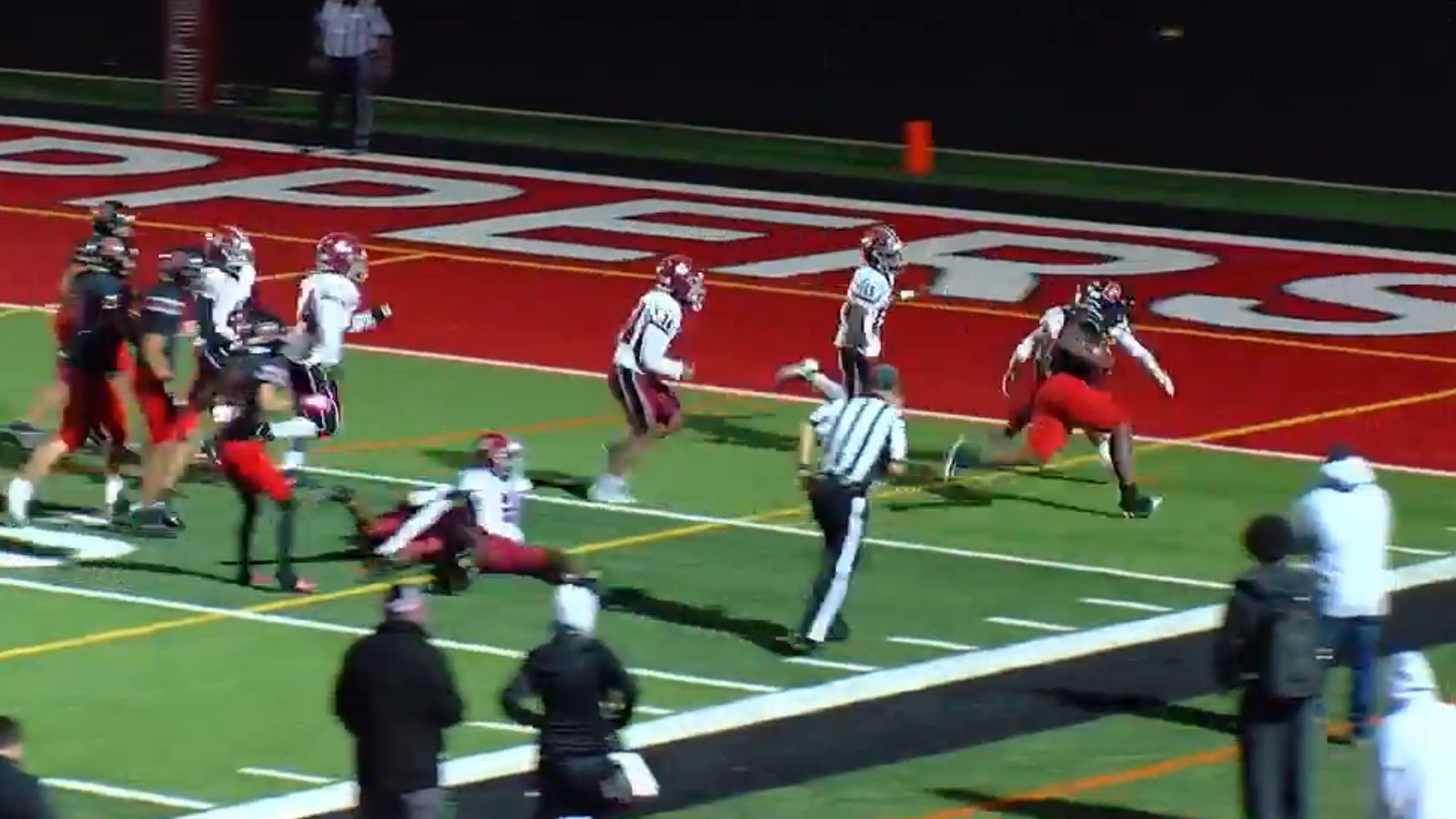 A Durfee ball carrier in a black jersey and red pants runs toward the corner of a red end zone lettered Hilltoppers as Brockton players in white and an official give chase, with fans along the sideline in the foreground