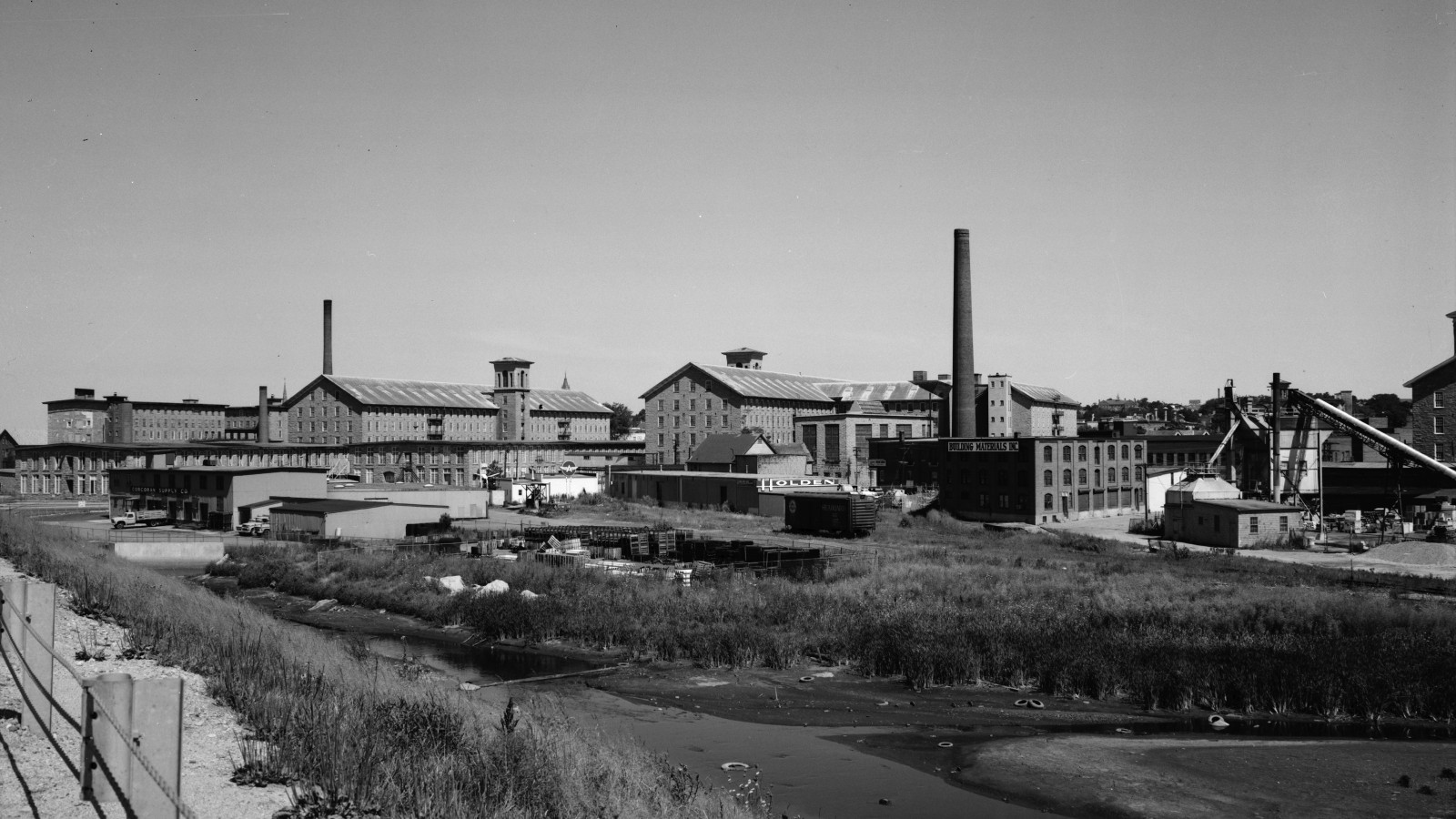 A black and white view of the Durfee Mills in Fall River in 1968: long granite mill buildings with towers and a tall smokestack behind a marshy stream bank and low industrial sheds