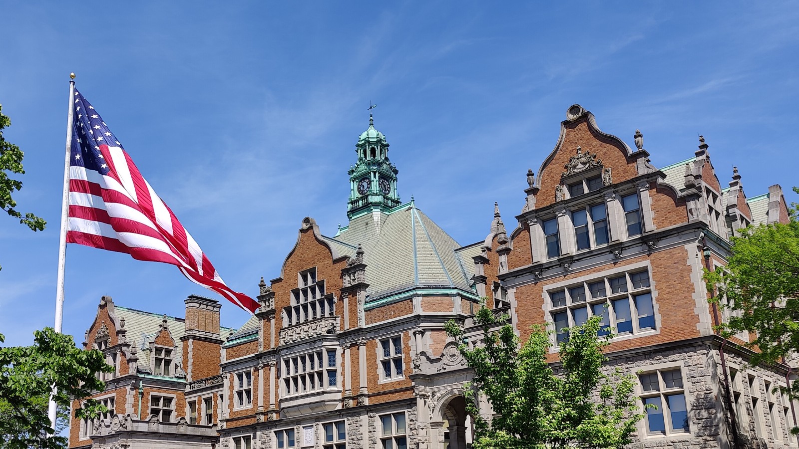 Fairhaven High School's brick and stone facade and green copper clock tower under a blue sky, with a large American flag flying from a tall pole at left