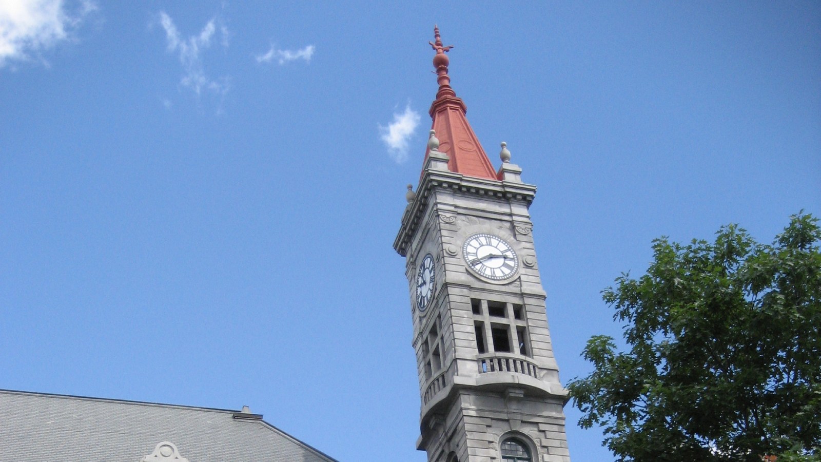 The granite clock tower of the 1886 B.M.C. Durfee High School building in Fall River, with two white clock faces and a red pointed roof, against a blue sky