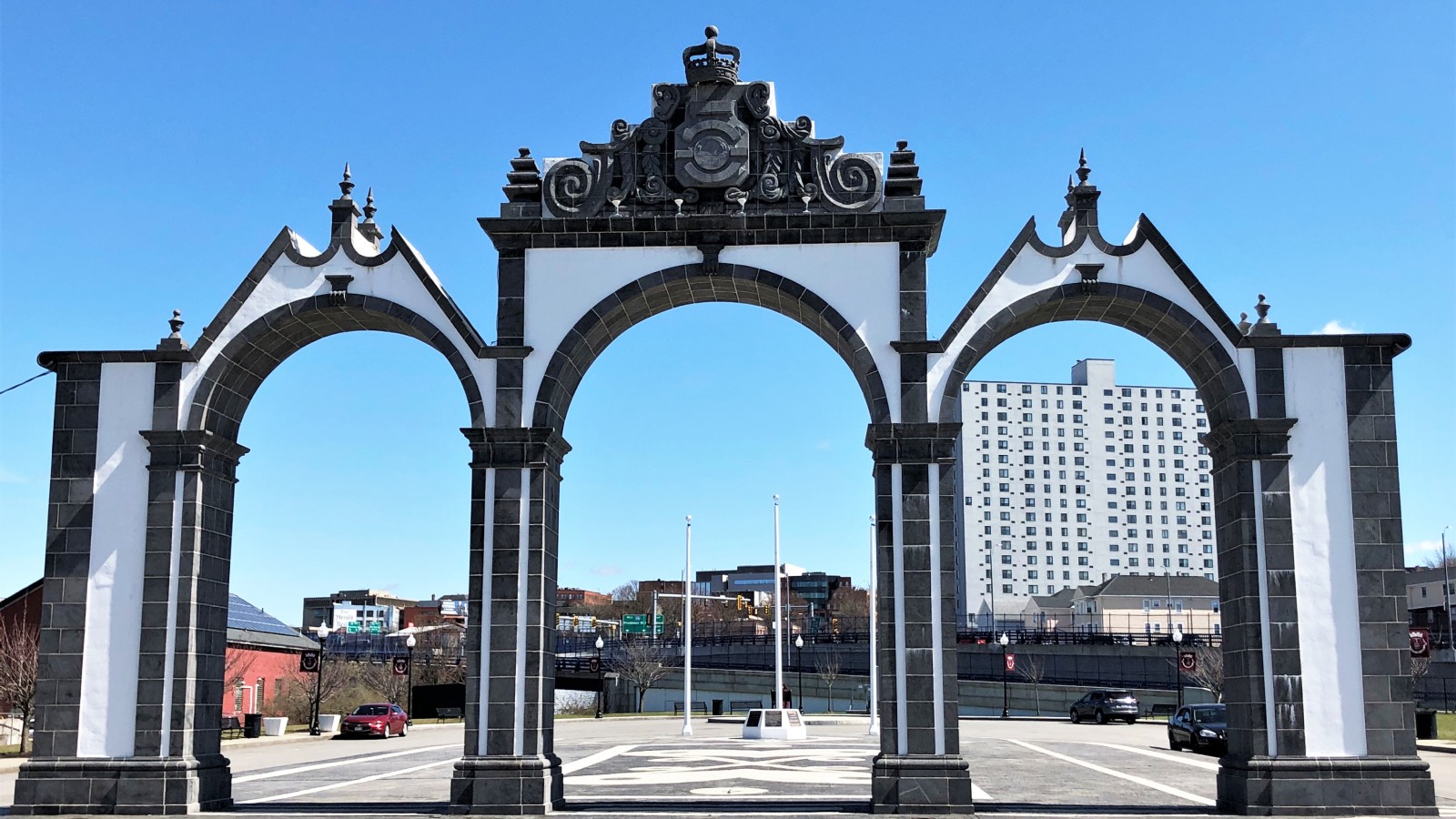 The white and dark gray stone arches of the Gates of the City monument on Fall River's waterfront, with parked cars, light poles and a high-rise behind them under a blue sky