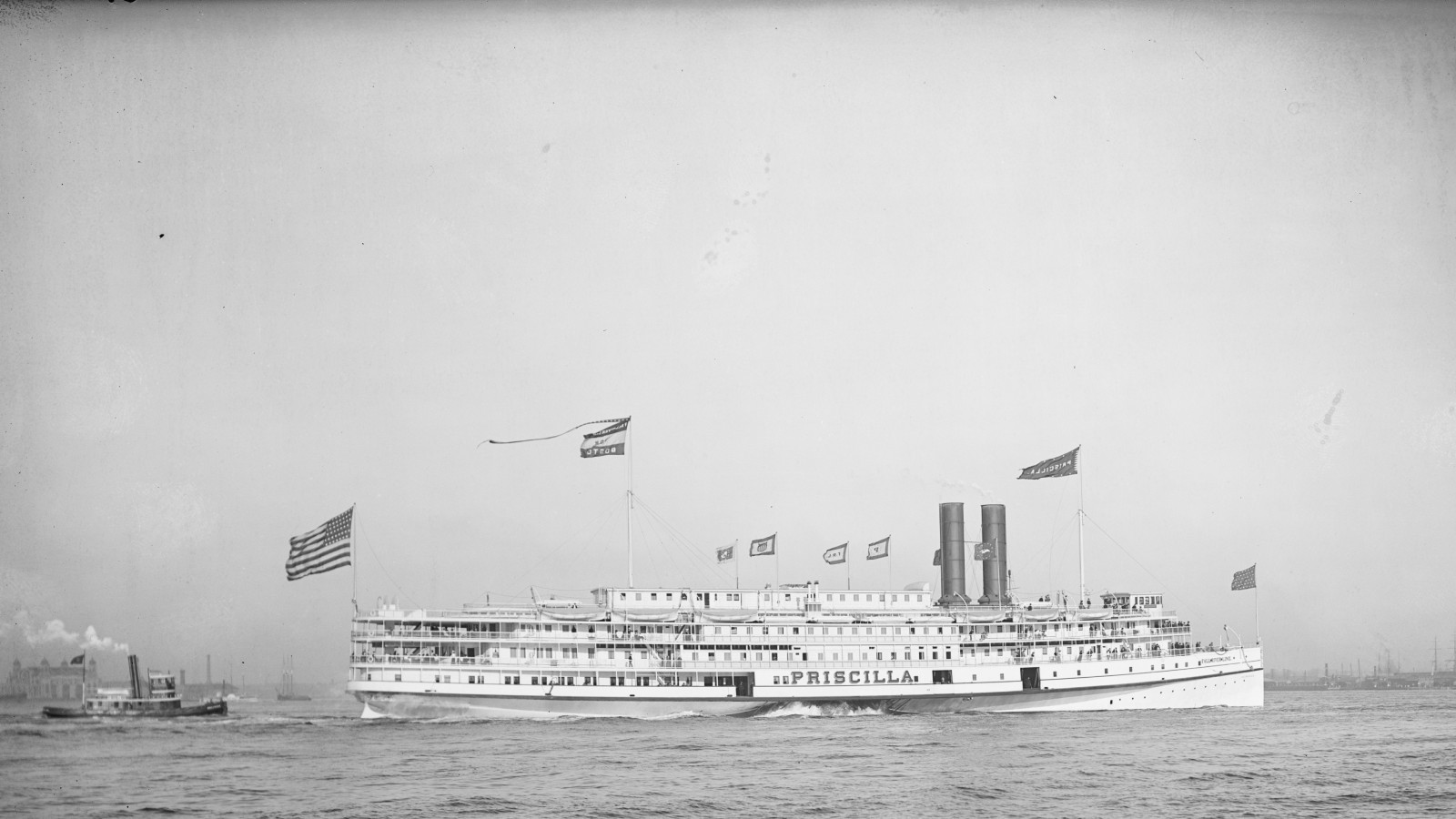 The white steamer Priscilla, flags flying, under way on open water with a tugboat behind her