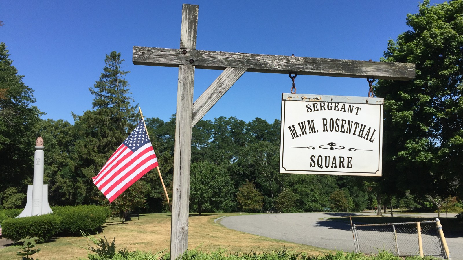 A white hanging sign reading Sergeant M.W.M. Rosenthal Square on a wooden post beside an American flag, with the lawns and trees of North Park and a park drive behind it