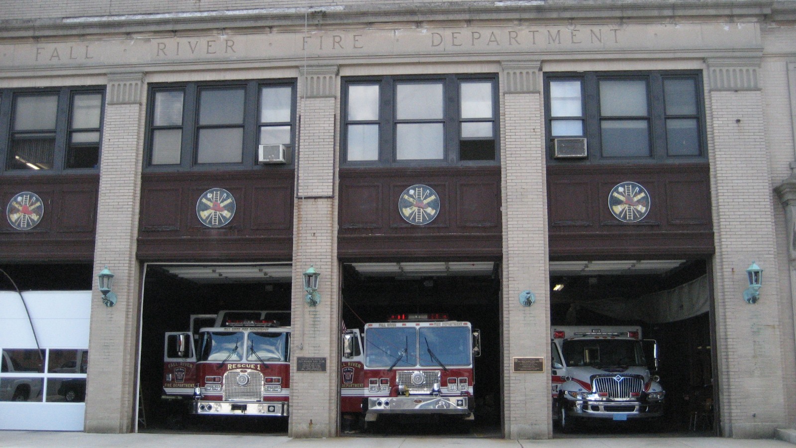 The open bays of Fall River's Central Fire Station, with a rescue truck and a fire engine in the left and center bays and an ambulance in the right bay, under the words Fall River Fire Department