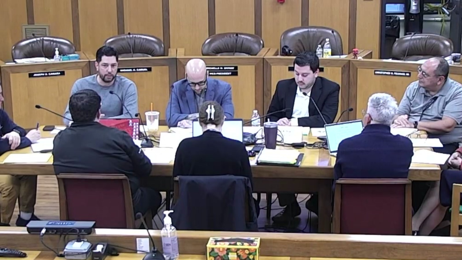 Members of a Fall River City Council committee sit at a long table in the council chamber with laptops and papers, facing school district officials whose backs are to the camera