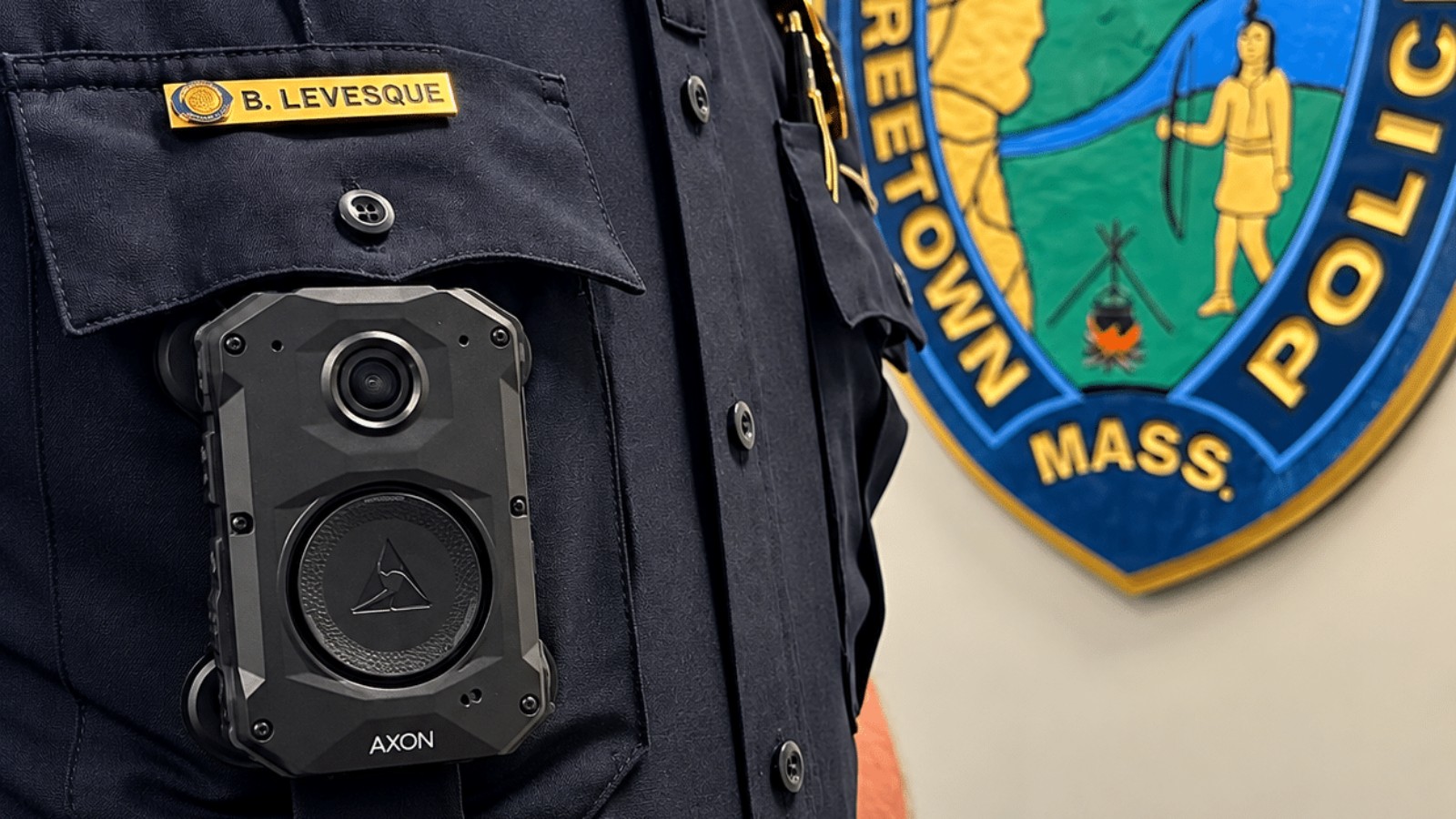 A Freetown police officer's uniform with an Axon body camera clipped to the chest pocket, the department's patch on the wall behind