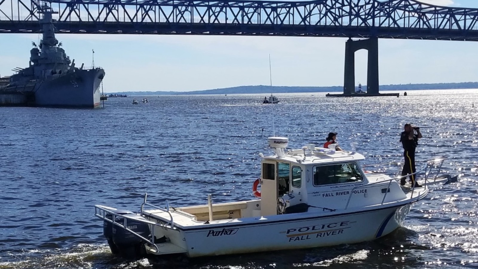 A white Fall River police boat marked POLICE FALL RIVER on the water, with an officer standing at the bow, the battleship USS Massachusetts at left and the steel Braga Bridge overhead