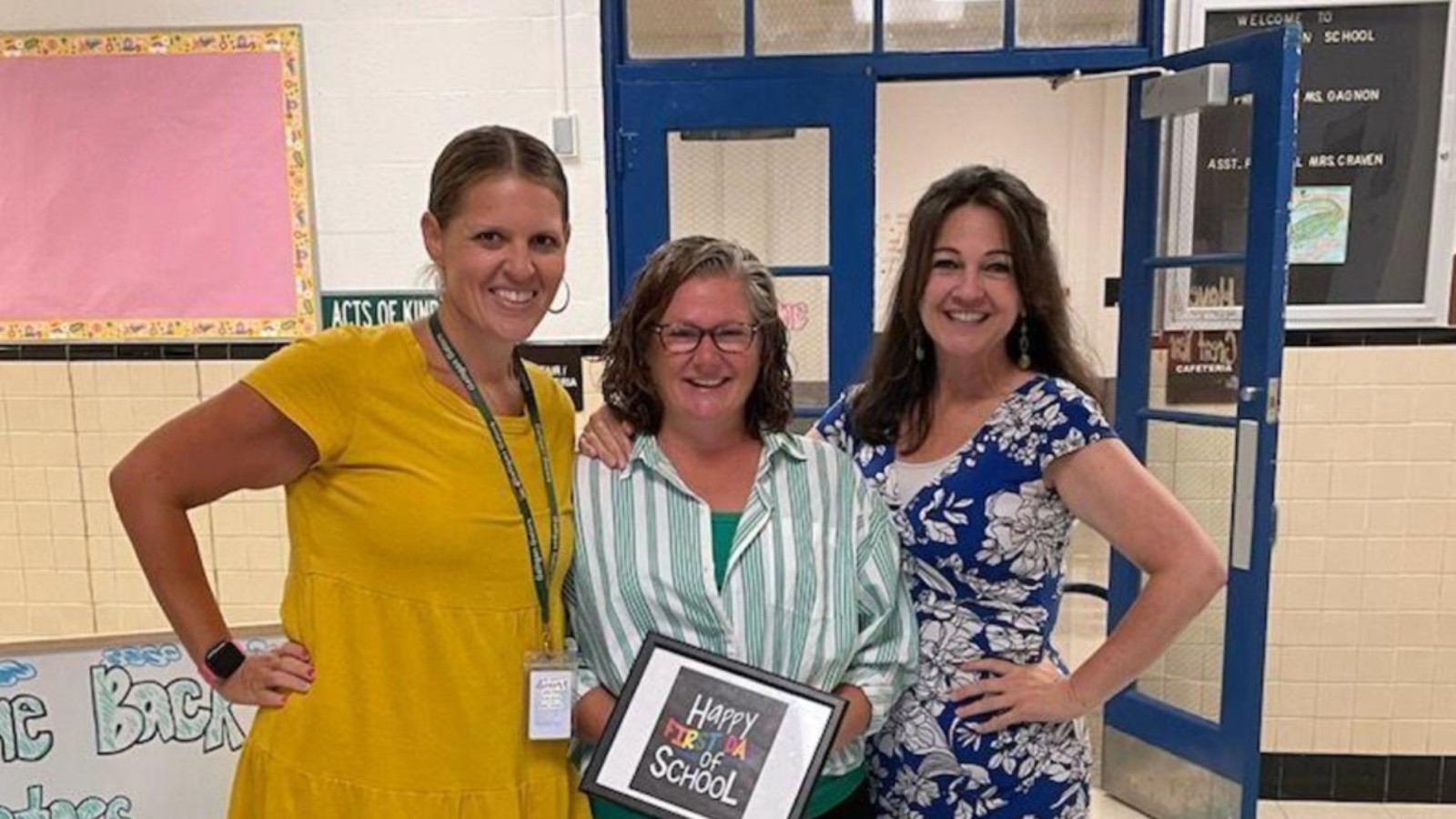 Three staff members smile in a school hallway in front of open blue doors, one holding a framed sign that reads Happy First Day of School, with a welcome board for Galligan School behind them