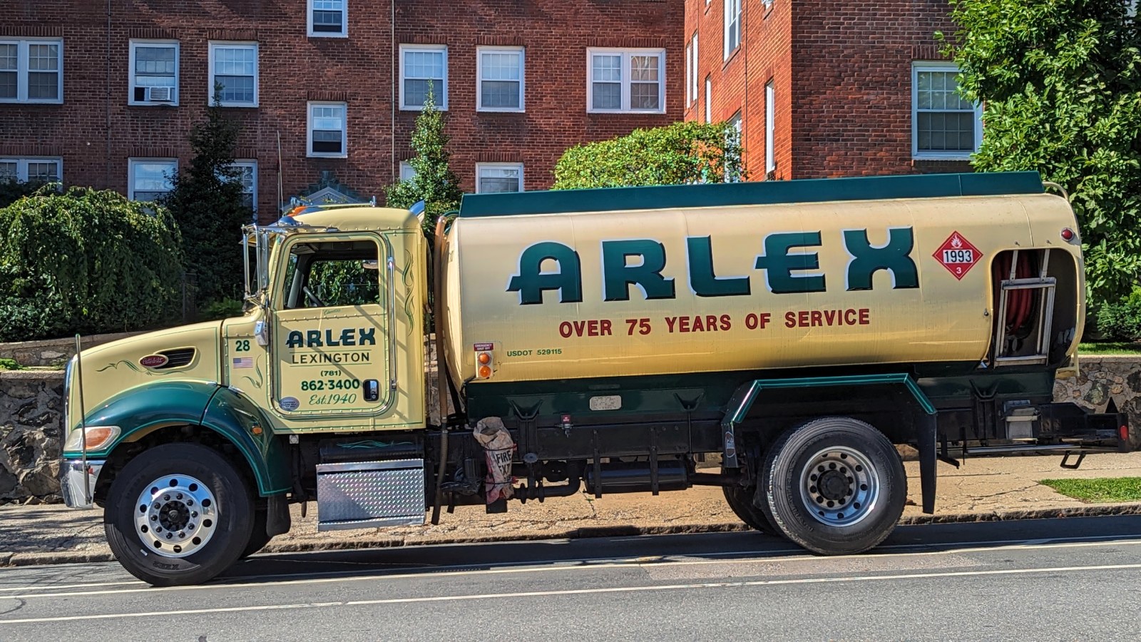 A cream and green home heating oil delivery truck with a red hazardous materials placard and a hose reel at the back, parked at the curb in front of red brick apartment buildings