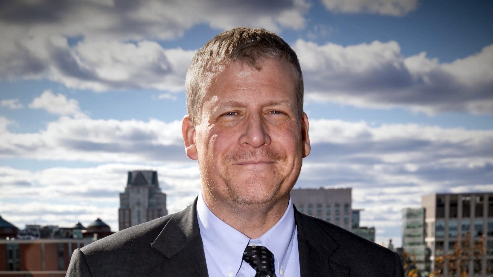Bristol County Sheriff Paul Heroux in a dark suit, white shirt and patterned tie, smiling outdoors with city buildings and a cloudy sky behind him