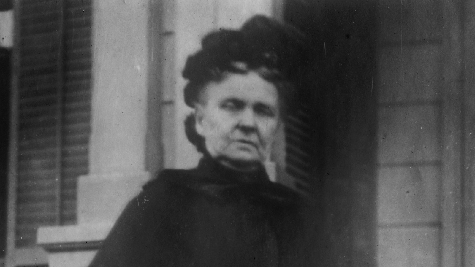 A black and white photo of Hetty Green, an older woman with dark hair piled up, in a black high-collared cape, standing outside a house