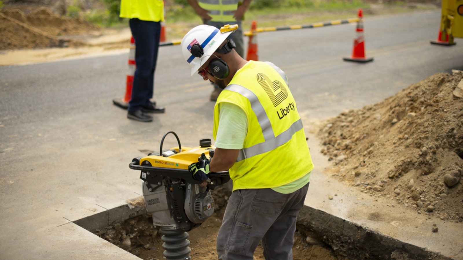 A Liberty gas worker in a white hard hat and a yellow Liberty safety vest runs a compactor in a trench cut into a paved road, with traffic cones and two crew members behind him