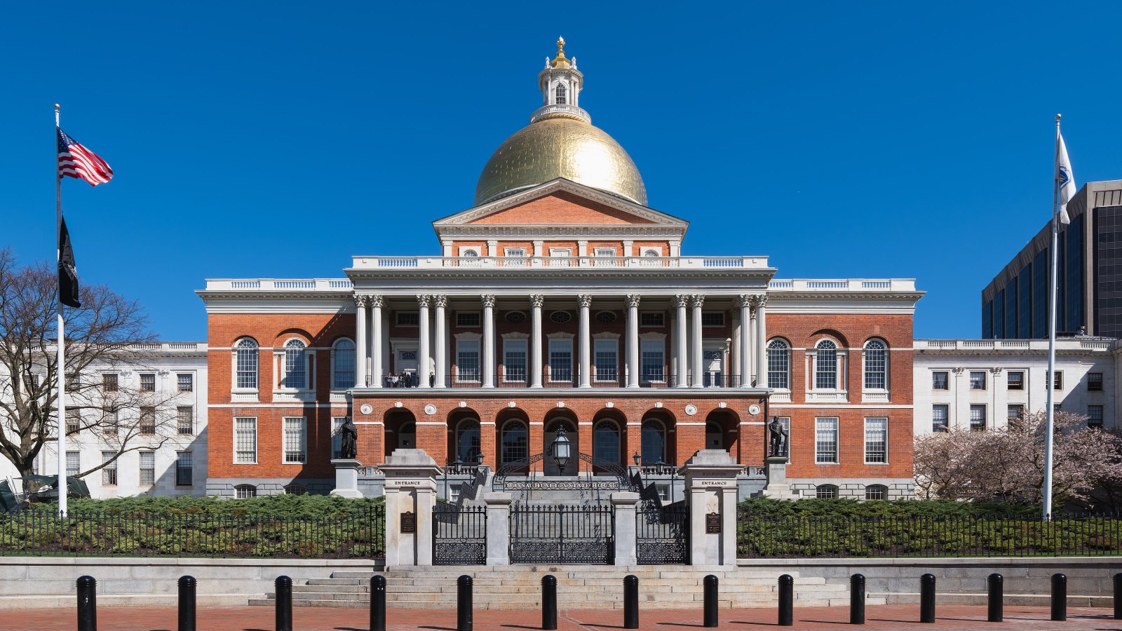 The Massachusetts State House in Boston, its gold dome over a red brick front with white columns, seen from Beacon Street behind its iron gates under a clear blue sky