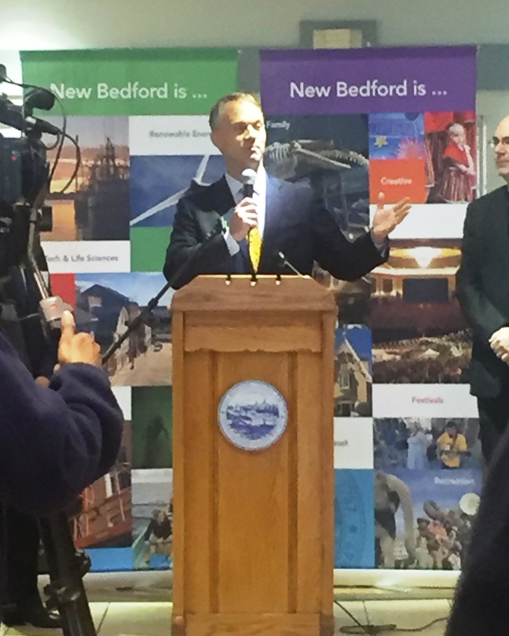 New Bedford Mayor Jon Mitchell speaking into a microphone at a wooden podium bearing the city seal