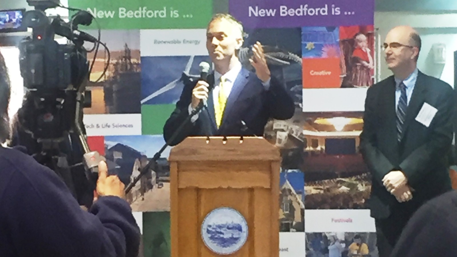 New Bedford Mayor Jon Mitchell speaking into a microphone at a wooden podium with the city seal, in front of New Bedford is banners