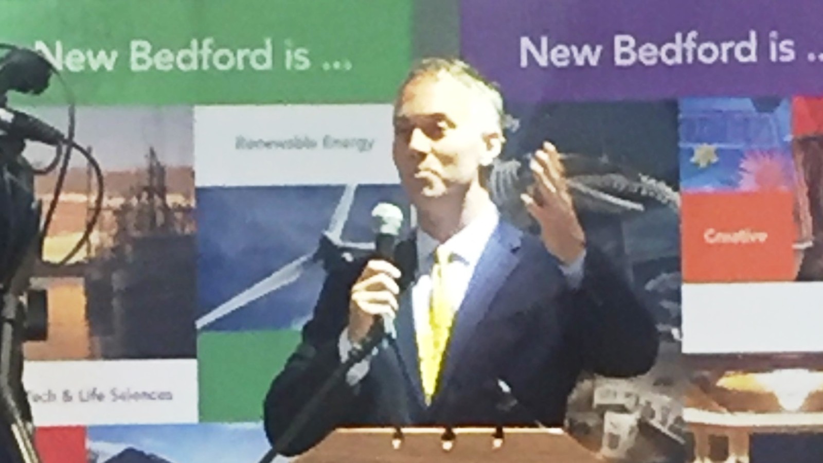 New Bedford Mayor Jon Mitchell speaking into a handheld microphone at a podium, in front of banners reading New Bedford is