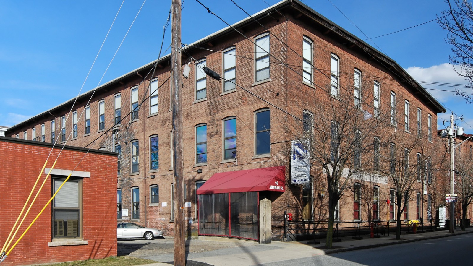 A three story brick mill building on Anawan Street in Fall River, with a red awning marked 16 Anawan St. over the entrance and a Narrows Center banner beside it