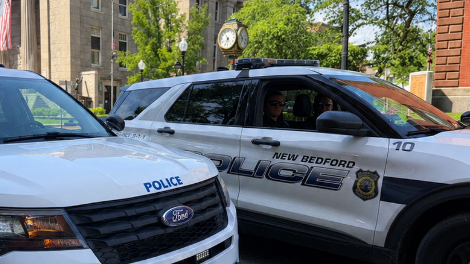 Two white New Bedford police SUVs parked side by side on a street, one marked POLICE on the hood and the other marked NEW BEDFORD POLICE with unit number 10 and two officers inside, with a stone building and a street clock behind them