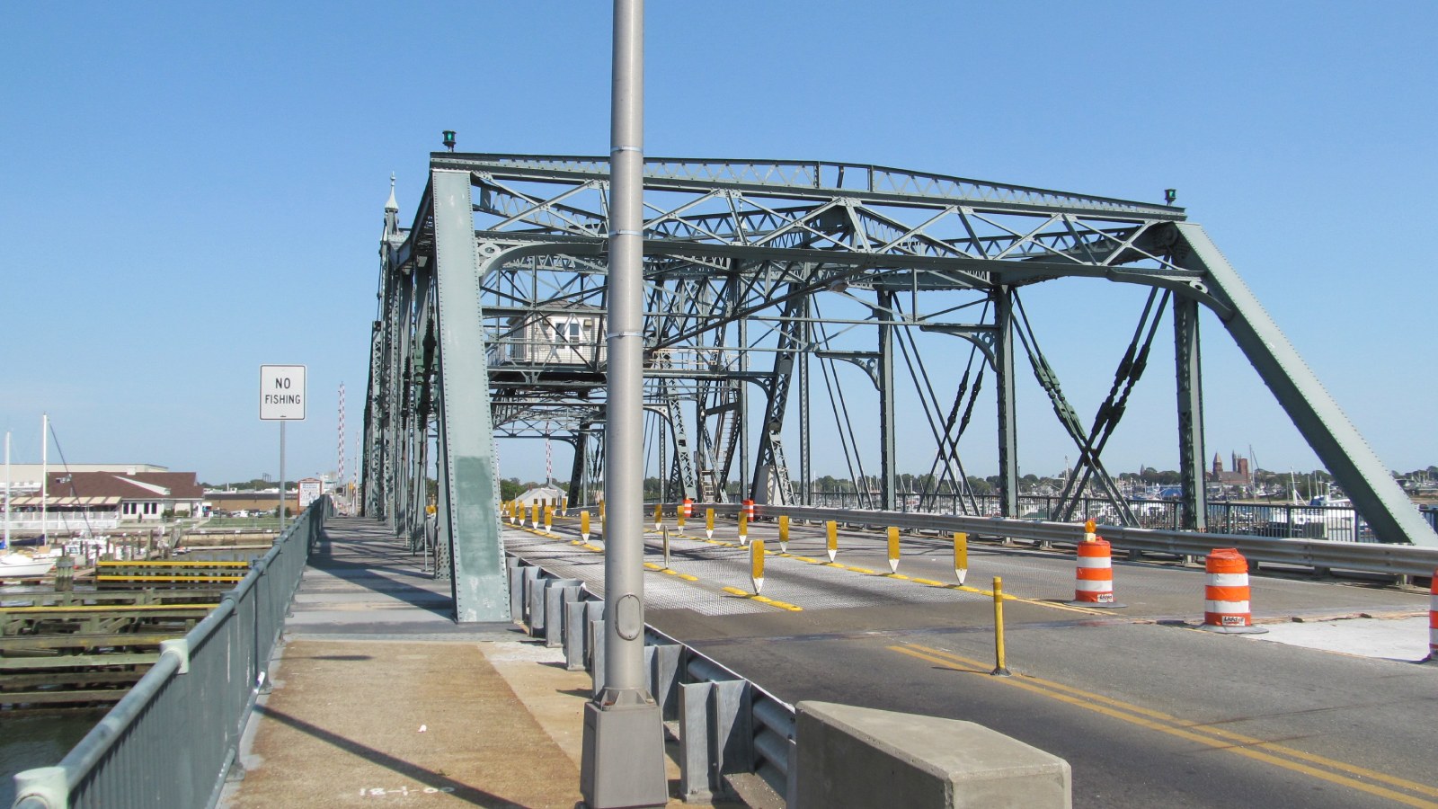 Looking along the roadway into the green steel truss of the New Bedford-Fairhaven swing span, with a small house on the span and boats in the harbor beyond
