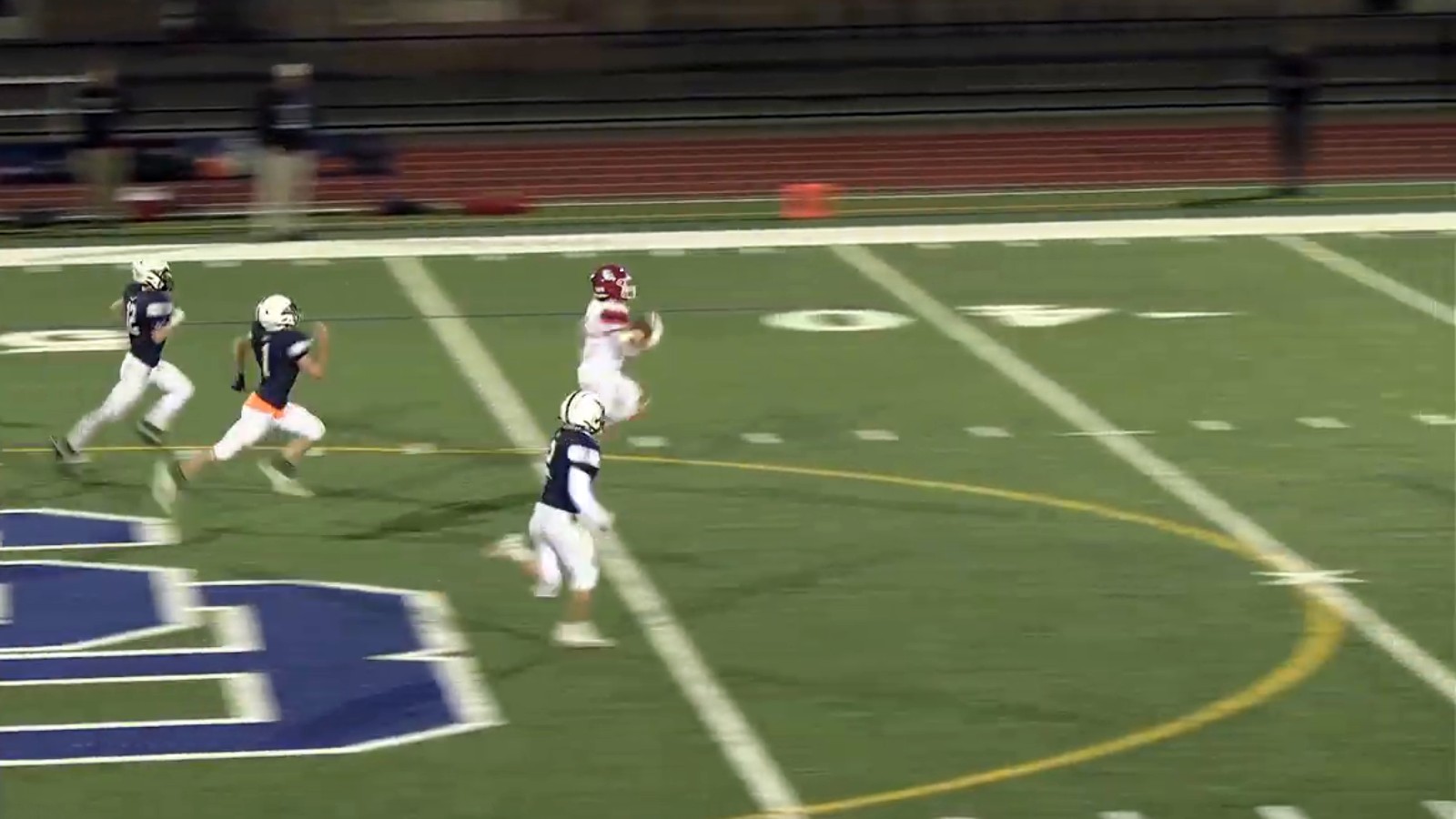 An Old Rochester ball carrier in a red helmet and white jersey runs clear across midfield as three Somerset Berkley players in navy jerseys chase him past the SB logo painted on the turf