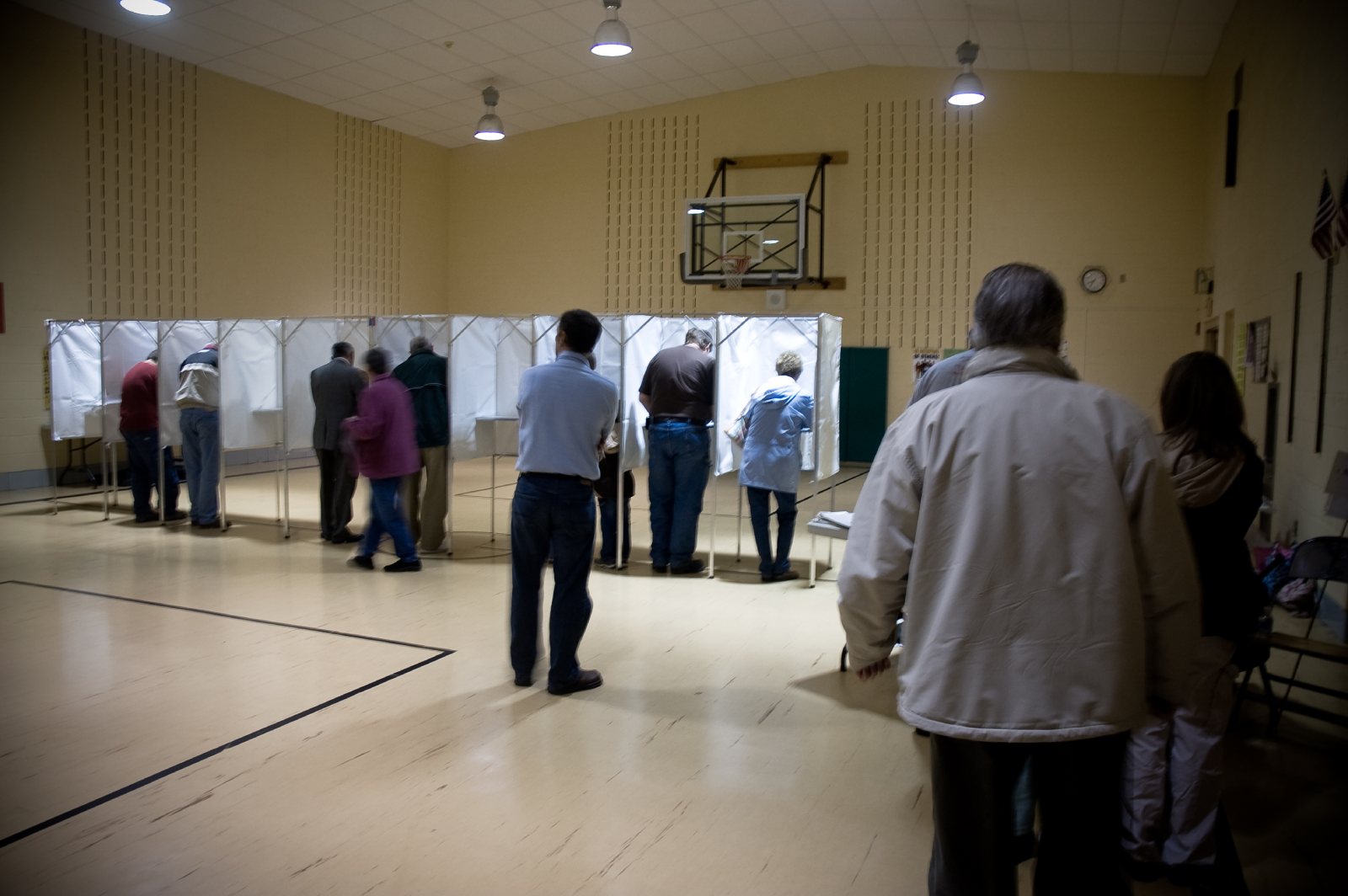 Voters standing at a row of white curtained voting booths in a school gym, with others waiting in line in the foreground