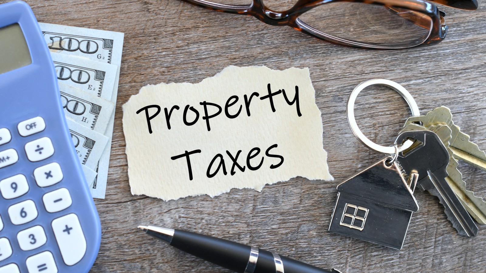 A torn paper note reading Property Taxes on a wood table, with a calculator, $100 bills, eyeglasses, a pen and house keys on a house-shaped key ring