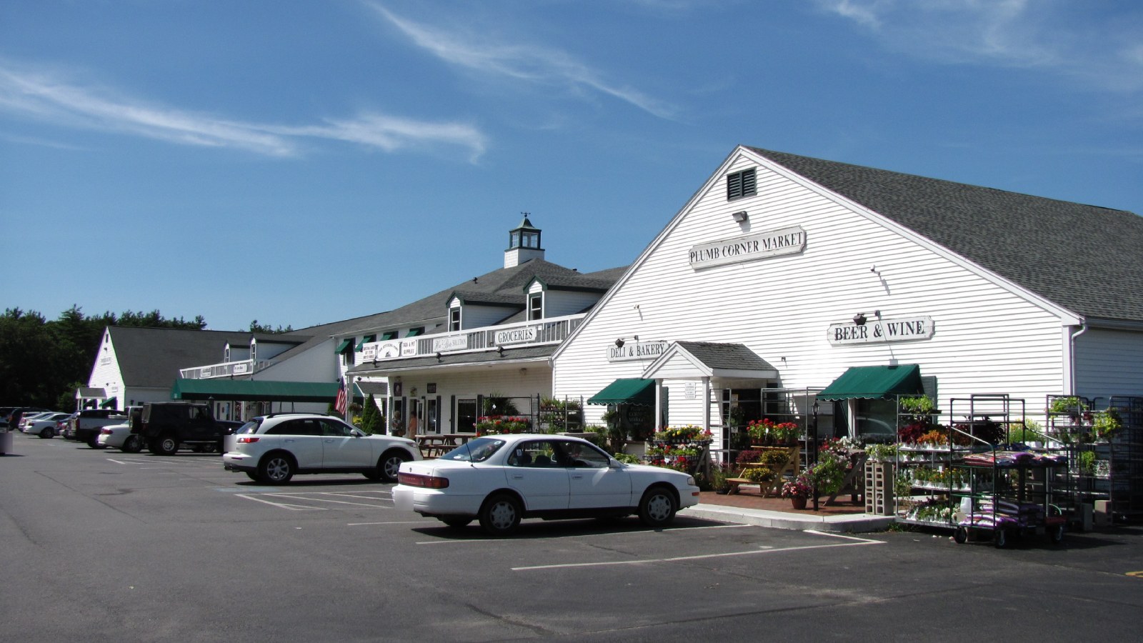 Plumb Corner Market in Rochester, a white clapboard building with green awnings and flower racks out front, beside a row of shops and a parking lot