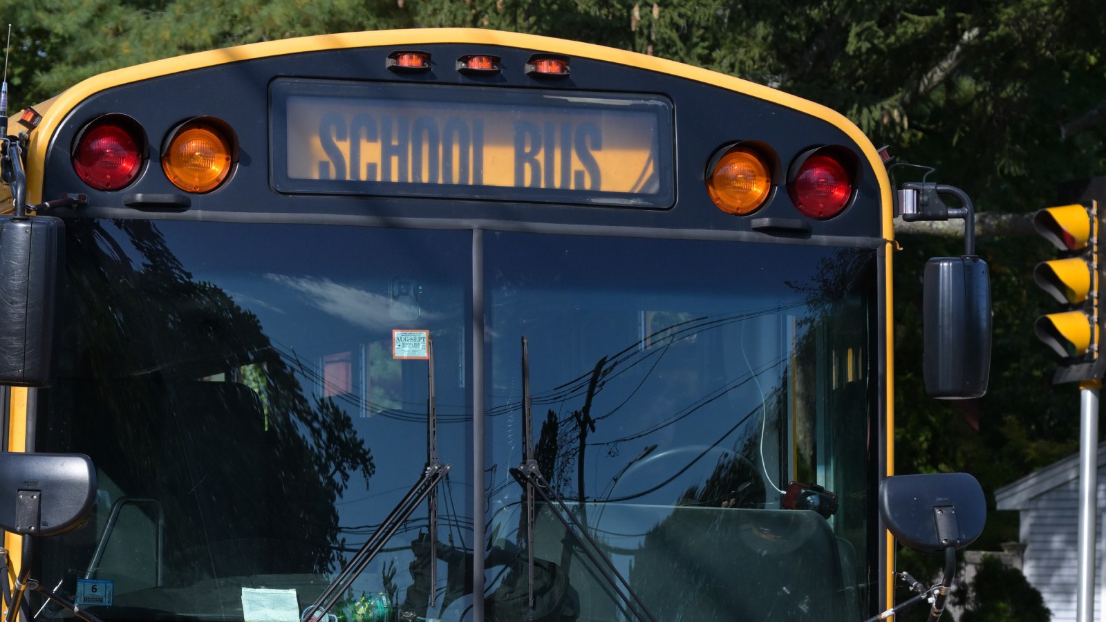 The front of a yellow school bus with a lit SCHOOL BUS sign above the windshield, red and amber warning lights and side mirrors, on a tree-lined street beside a traffic signal
