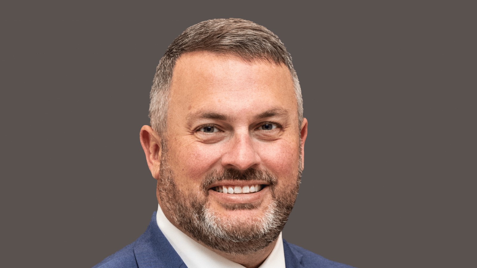 Stephen Canessa, smiling, with short graying hair and a beard, in a blue suit jacket, white shirt and patterned tie against a plain gray backdrop