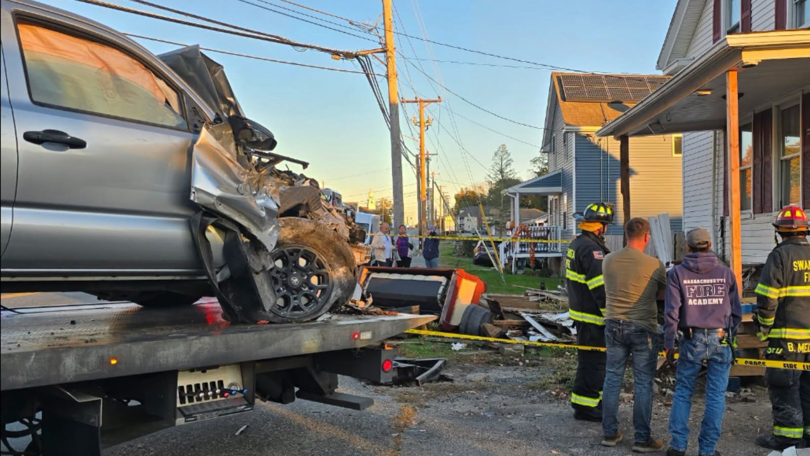 Firefighters stand by a damaged porch as a gray pickup truck sits on a flatbed tow truck after a crash into a Swansea home