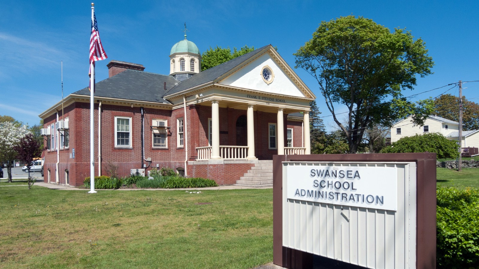 A red brick school building with a white columned porch, a green domed cupola and Frank S. Stevens School over the entrance, behind a sign reading Swansea School Administration