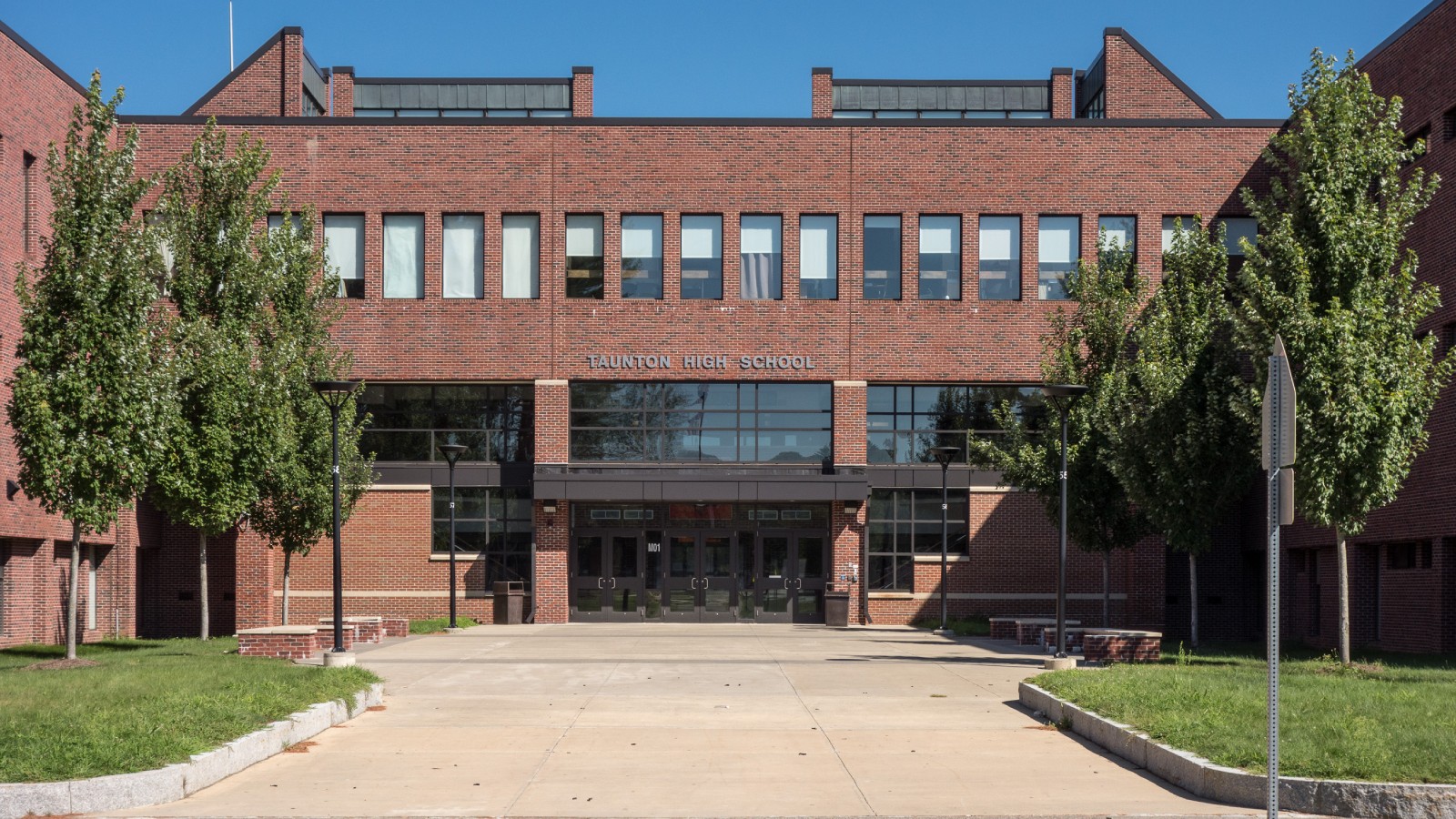 The brick front of Taunton High School with its name above a glass entrance, young trees on both sides and a wide paved walk leading to the doors