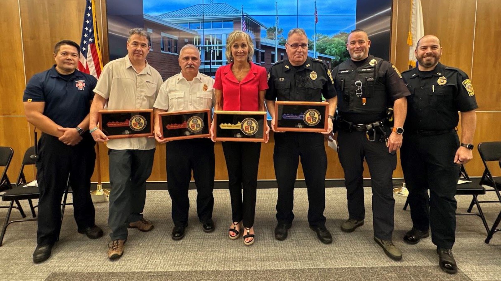 Seven people stand in a row in a meeting room at Taunton's new Public Safety Facility, four of them holding framed ceremonial key plaques, several in police and fire uniforms, in front of a video wall showing the building
