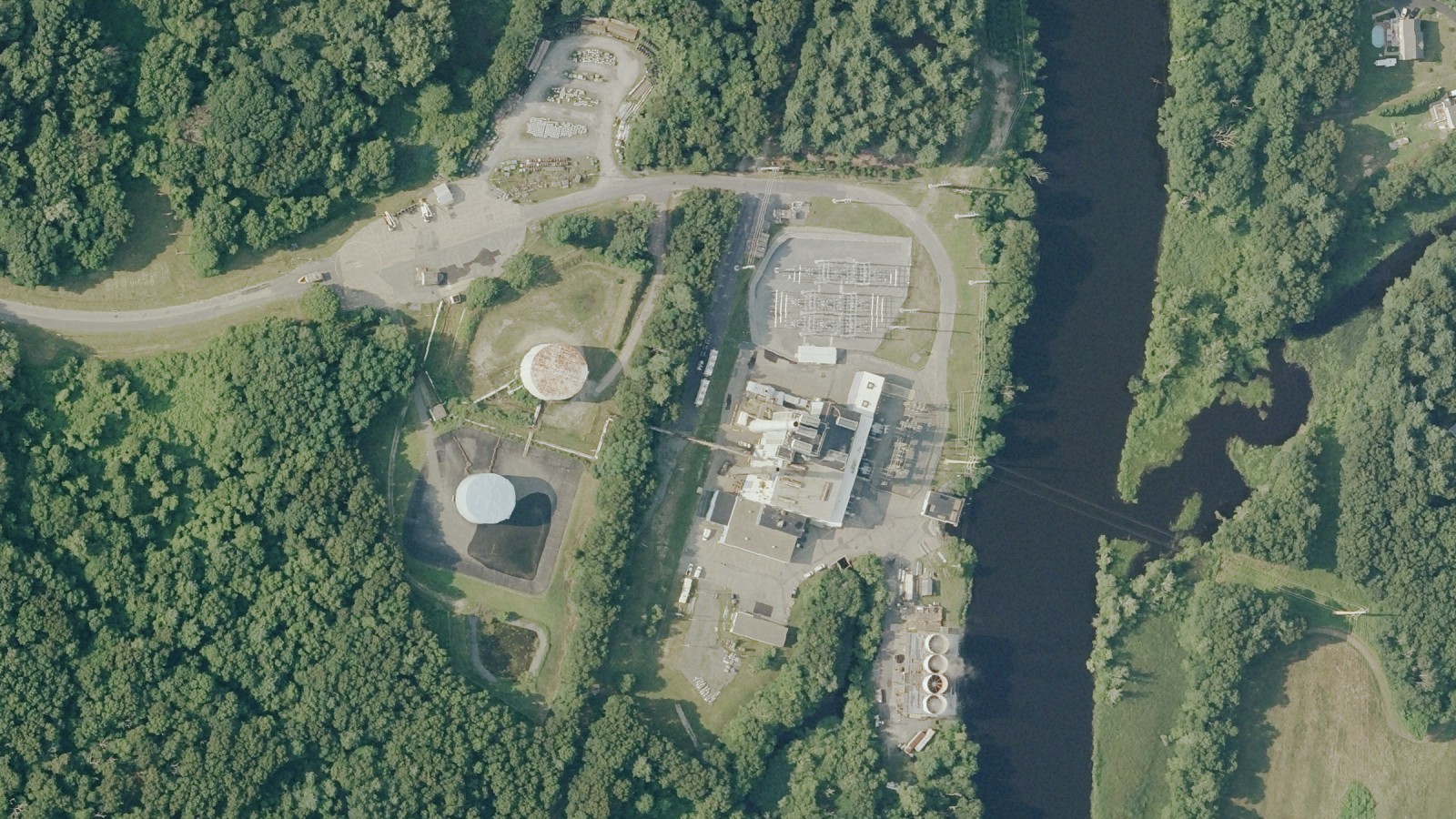 Aerial view of the Taunton Municipal Lighting Plant's Cleary-Flood Generating Station in Taunton: the white generating building and its fenced switchyard at center, two round tanks at left and a river along the right, surrounded by woods