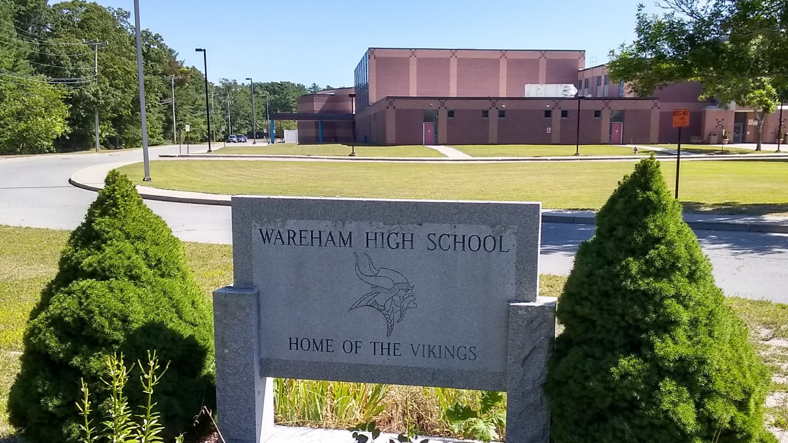 A granite sign reading Wareham High School, Home of the Vikings, between two shrubs, with the brick school building across a lawn behind it