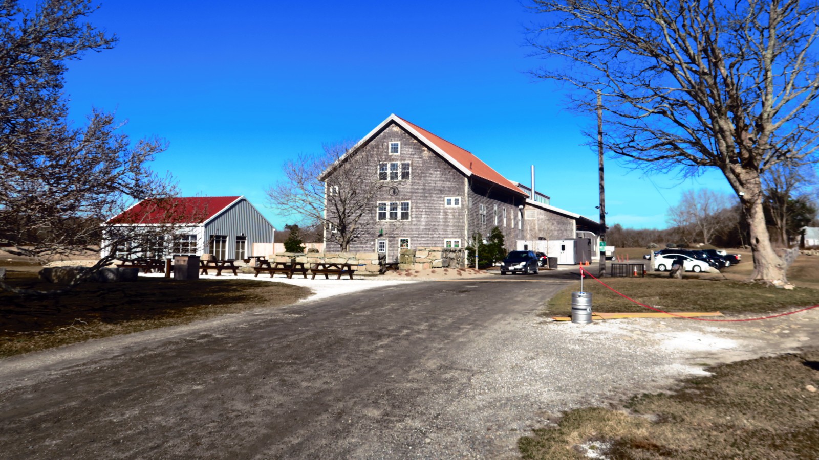 Buzzards Bay Brewing's tall gray shingled building with a rust-red roof, beside a smaller gray building with picnic tables and a gravel drive, under a clear blue sky in Westport