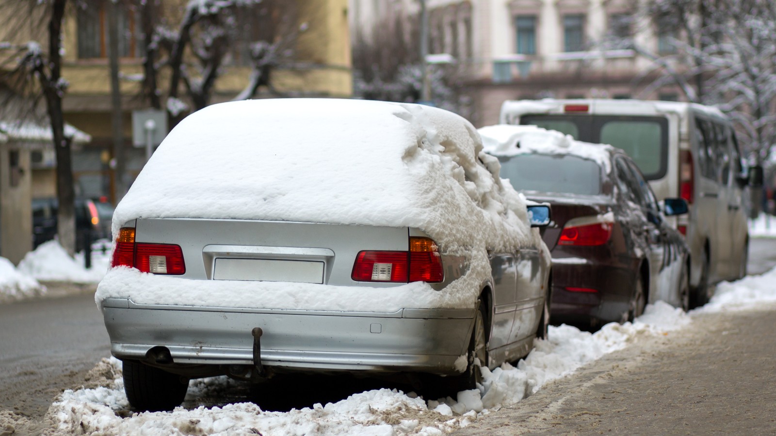Snow-covered cars parked along a slushy city street curb